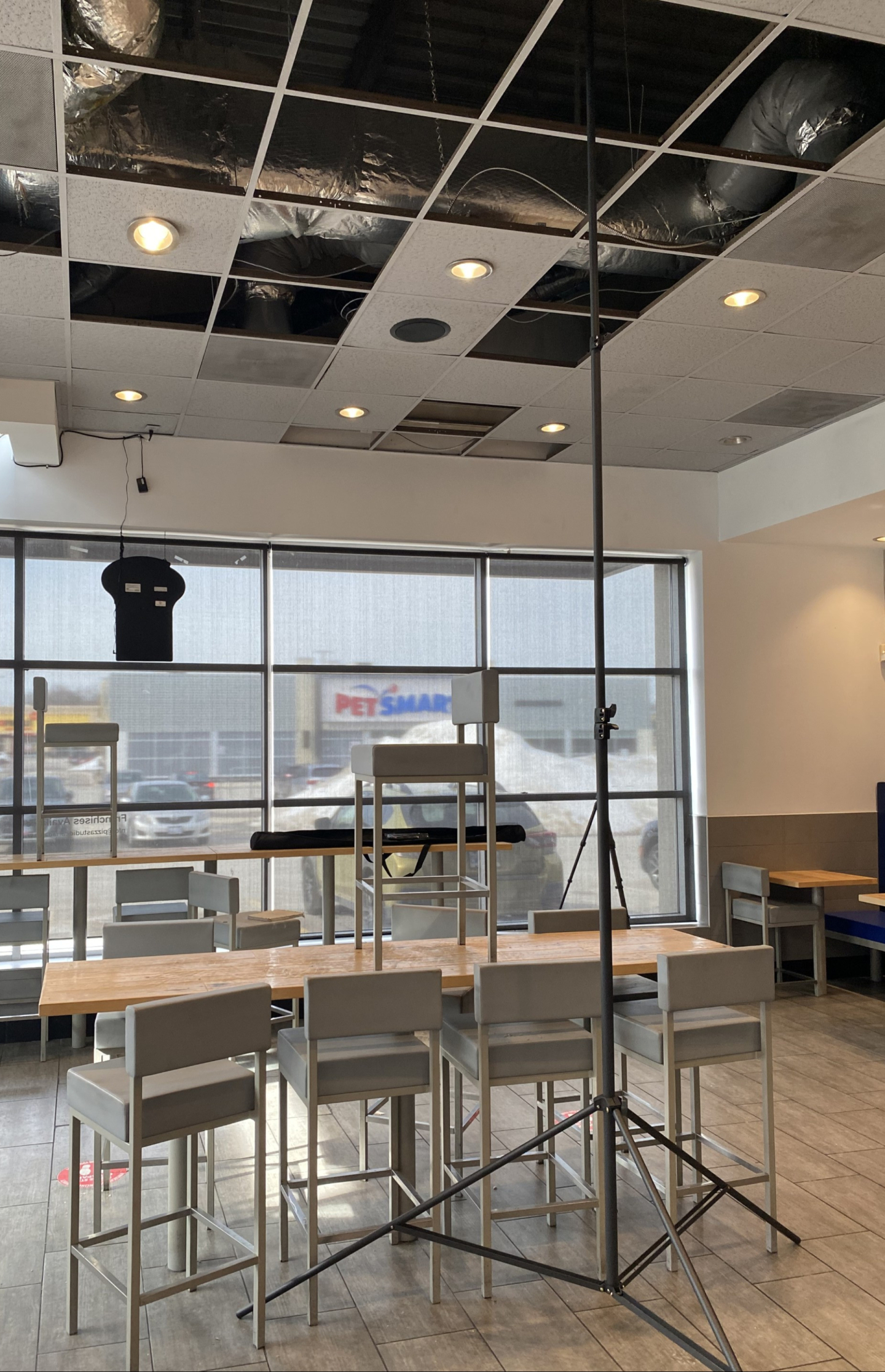 An interior dining area with high tables and stools arranged near large front windows. A lighting stand and equipment are set up beneath a partially open drop ceiling with exposed ductwork and recessed lights.